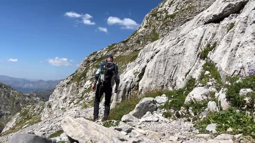 A Man with a Backpack Walks Along a Mountain Slope Surrounded By Majestic Mountains Rear View
