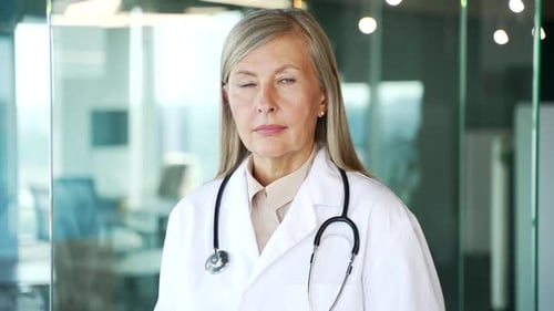 Portrait of serious senior gray haired female doctor in white coat looking at camera in hospital