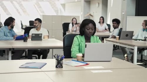 Jeune femme d'affaires africaine concentrée travaillant à son bureau sur un ordinateur portable dans un bureau moderne avec des collègues