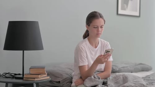 Young Woman Uses Phone Relaxing on Bed Indoors