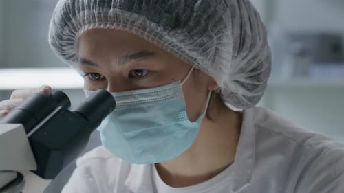 Asian Female Scientist in Mask Observing Sample through Microscope