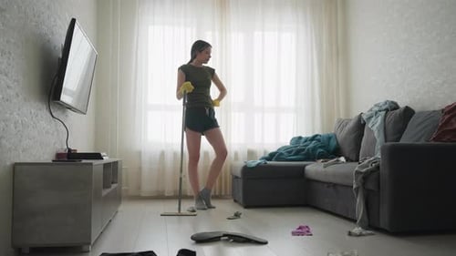 Lady Standing in Messy Living Room with Mop and Gloves
