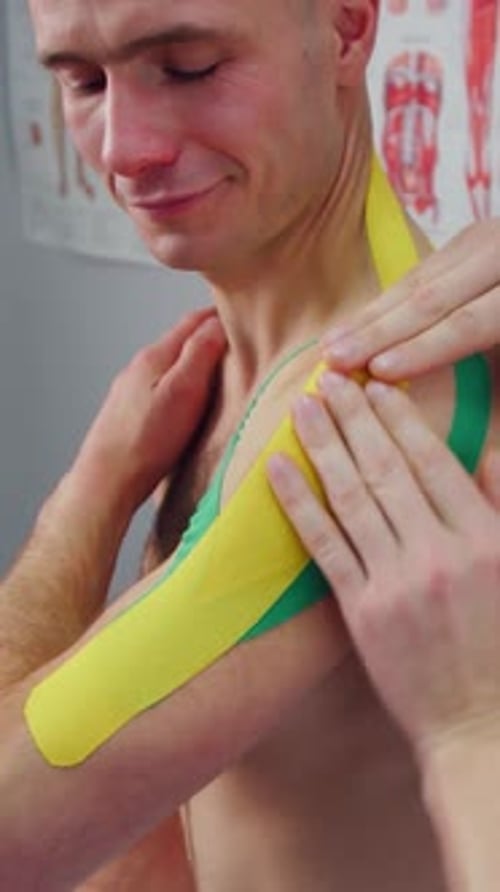 Physiotherapist Doing Kinesio Taping on the Shoulder of a Young Man in Clinic A Doctor Applies