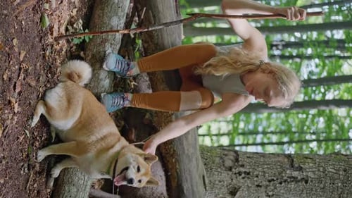 a Blonde Woman Sitting on a Fallen Tree in the Forest Petting the Dog Shiba Inu