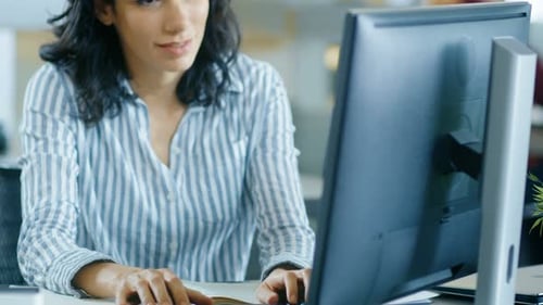 Beautiful Young Hispanic Woman sits at Her Desk and Works on a Personal Computer. In the Backgroun