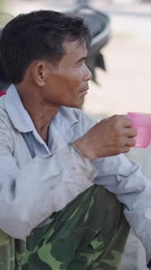 Asian Mature Man Squatting in a Very Dirty Shirt Drinks Water From a Portable Cooler in Vietnam
