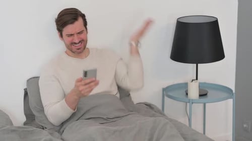 Young Man Upset by Loss on Phone while Sitting in Bed
