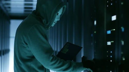 Masked Hacker wearing Hoodie Opens Server Cabinet and Connects to Server with His Notebook.