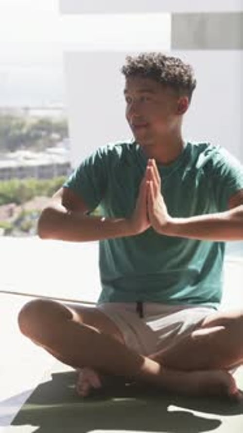 Camera panning, man moving into seated cross-legged pose on yoga mat, holding palms for wellness