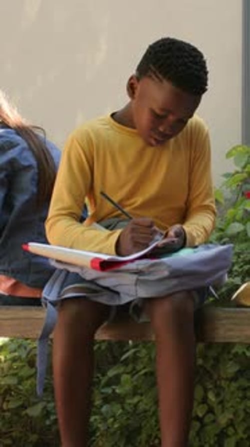 Vertical video: Leaning youth male writing with pencil on notebook on bench at school in yellow top