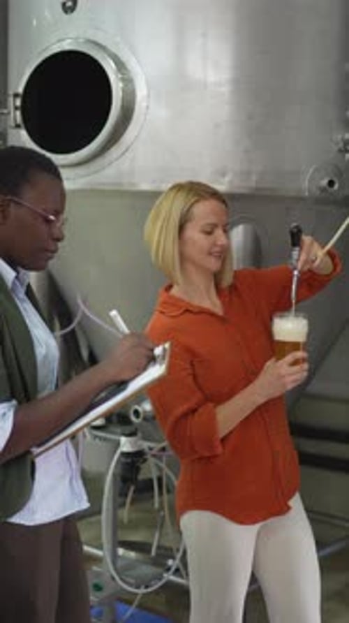 Two female brewers conducting quality control by pouring and examining fresh craft beer from a tank
