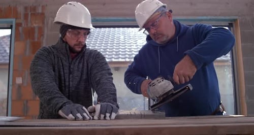 Two construction workers using angle grinder in workshop, wearing hard hats and safety gear, sparks