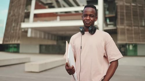 Young student stands outside of university wearing headphones, smiling and looking at the camera