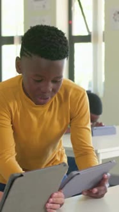 Vertical video: Walking youth in yellow shirt leaning at class desk, comparing two covered tablets