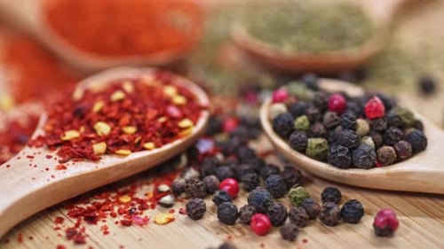 A close-up of various spices on wooden spoons, featuring crushed red pepper flakes and a mix of blac