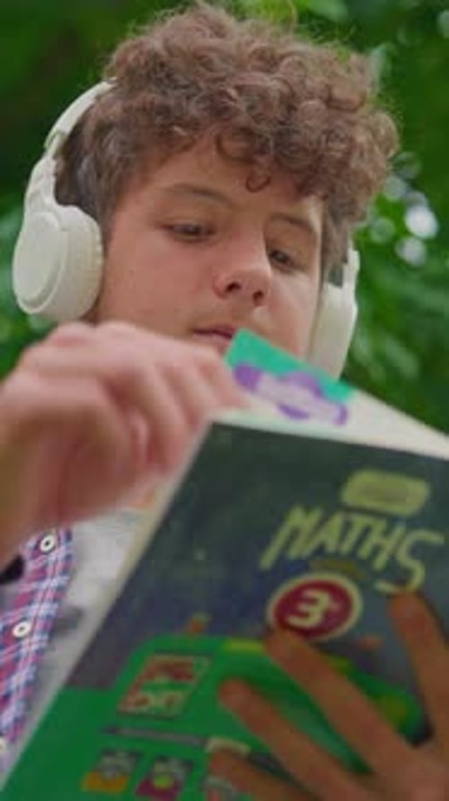 Vertical Video Curly Teenage Boy Listening to Music on Headphones and Reading a Book While Sitting