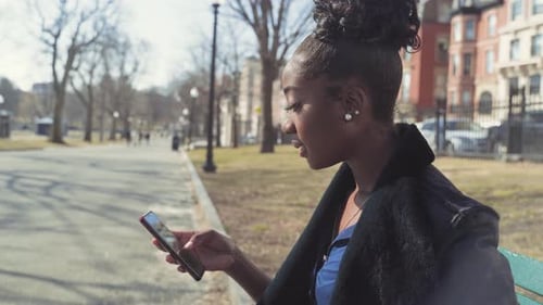 Pretty Afro-American Girl Laughing While Browsing On Her Phone In The Park, Sitting On The Bench On
