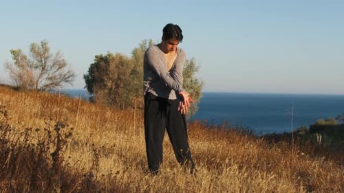 Young Man Stretching by Ocean on Grassy Hillside