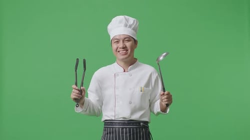 Asian Man Chef Smiling And Showing Tongs And Ladle To Camera While Standing In The Green Screen