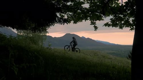 Athletic man pedalling a MTB E-bike up a steep grassy hill. Beautiful view of the mountains.
