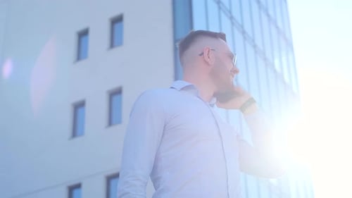 A Man with a Beard Wearing a Dress Shirt Stands in Front of the Building Talking on His Cell Phone