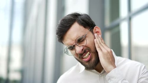 Businessman suffers from ear pain standing on street near an office building. Man touches his ear