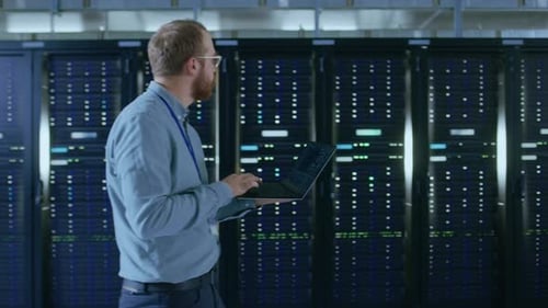 Bearded Data Center IT Engineer in Glasses is Walking Through Server Rack Corridor with a Laptop Co