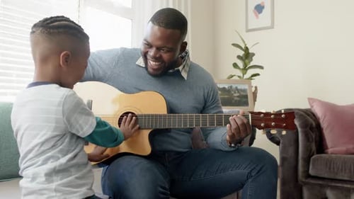 Happy african american father and son sitting on sofa and playing guitar, in slow motion