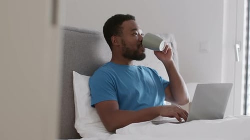 Man Works on Laptop in Bed and Drinks
