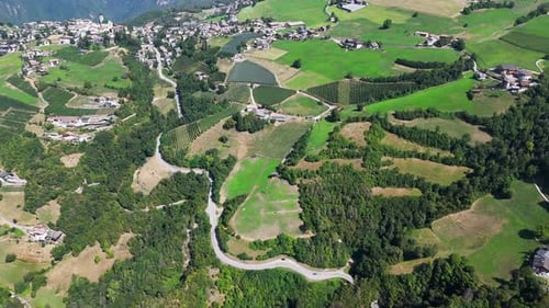 Camera pans over a winding road as it cuts through a mountain village and surrounding green valley