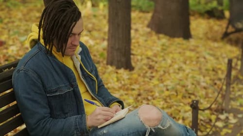 Young Creative Man with Dreadlocks Drawing Writing in Notebook Sitting on Park Bench in Golden