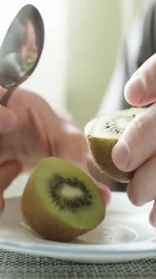 A man cuts and eats a kiwi fruit with a spoon, in the sun, close-up. Vertical video.