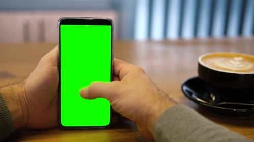 Young Man Sitting at Cafe Holding Smartphone Green Mockup Screen in Hand Male Person Using Chroma