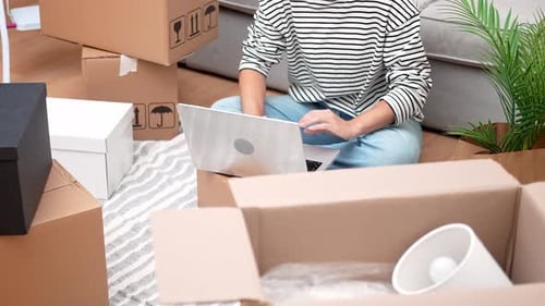 Woman Surrounded By Boxes Uses a Laptop to Plan Home Decor Shop Online or Book Removal Services