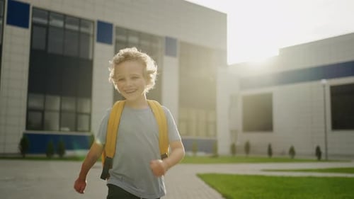 Porträt eines fröhlich lächelnden Jungen mit Rucksack, der aus dem Schulgebäude rennt, Zeitlupe, mittlere Brennweite