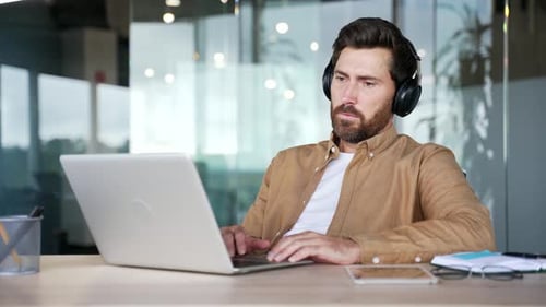 Happy relaxed IT specialist in wireless headphones works on a laptop sitting at a workplace
