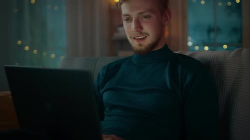 In the Evening Handsome Young Man at Home Sitting on a Couch in the Living Room Works on a Laptop C