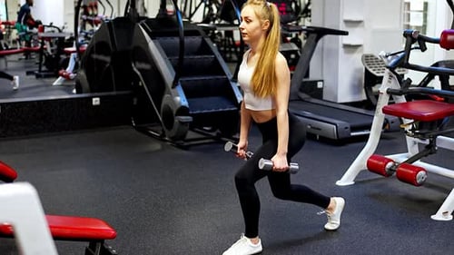 Young Woman Holds Dumbbells in Her Hands and Works Out in the Gym Performing an Exercise