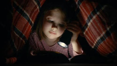 Smart Little Girl Reads Interesting Book with a Flashlight Under Blanket.