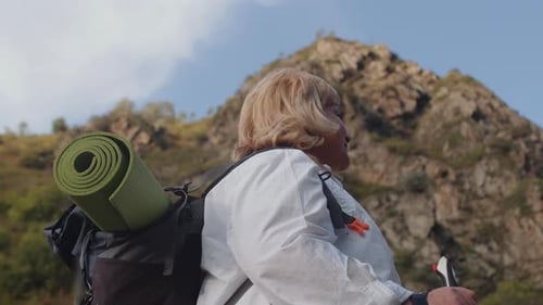 Elderly Woman with a Backpack and Hiking Sticks Looking Around and Continuing Her Mountain Walk