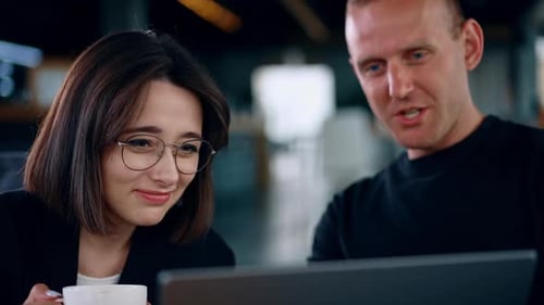 Man and woman look at laptop screen laughing. Lady wearing glasses holds a cup in hand. Close up.