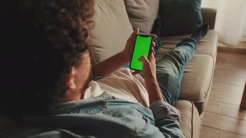 Young man lying on sofa uses mobile phone with green mock-up screen chroma key