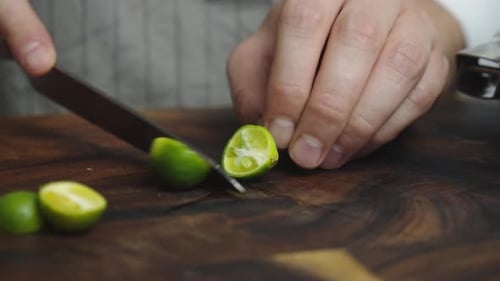 Slicing green lime lemon with kitchen knife on wooden cut board. Close up shot. Slow motion.