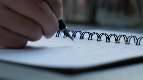 The male is writing on a white textbook with a pen on the desk.