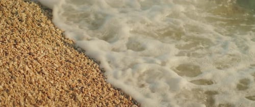 Foamy waves reach pebble beach on shore under the golden sunlight close up