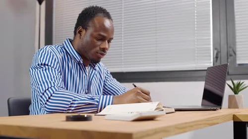 Serious young African businessman works with documents, looks at his laptop and writes in a notebook