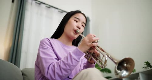 A young woman practices playing the trumpet while sitting on a sofa in a cozy living room.