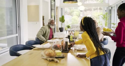 Diverse family greeting, hugging, pulling chairs, arranging plates for meal at home dining table