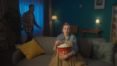 Shocked Woman Watching a Scary Movie on TV at Home and Eating Popcorn A Man Comes Up to a Woman From