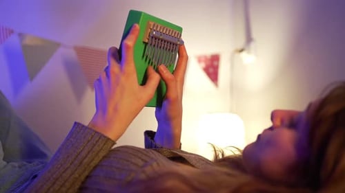 Woman Relaxing on Bed Softly Illuminated Playing Green Kalimba Generating Serene Musical Ambiance